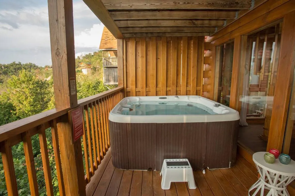 The image showcases a hot tub set on a wooden deck, bordered by a railing and surrounded by green trees. The tranquil outdoor ambiance offers a glimpse of the nature views guests can enjoy while soaking in the tub, suggesting post-adventure relaxation and group bonding.