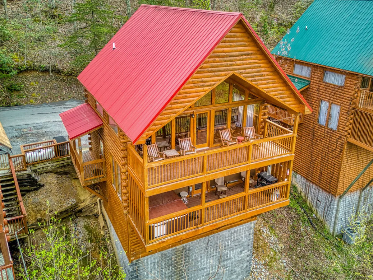 A classic log cabin with a red roof stands amidst trees, featuring a spacious wooden deck with rocking chairs. The setting feels both cozy and adventurous, hinting at the privacy and charm of cabin life in the Smokies, where families can unwind after exploring nearby attractions.