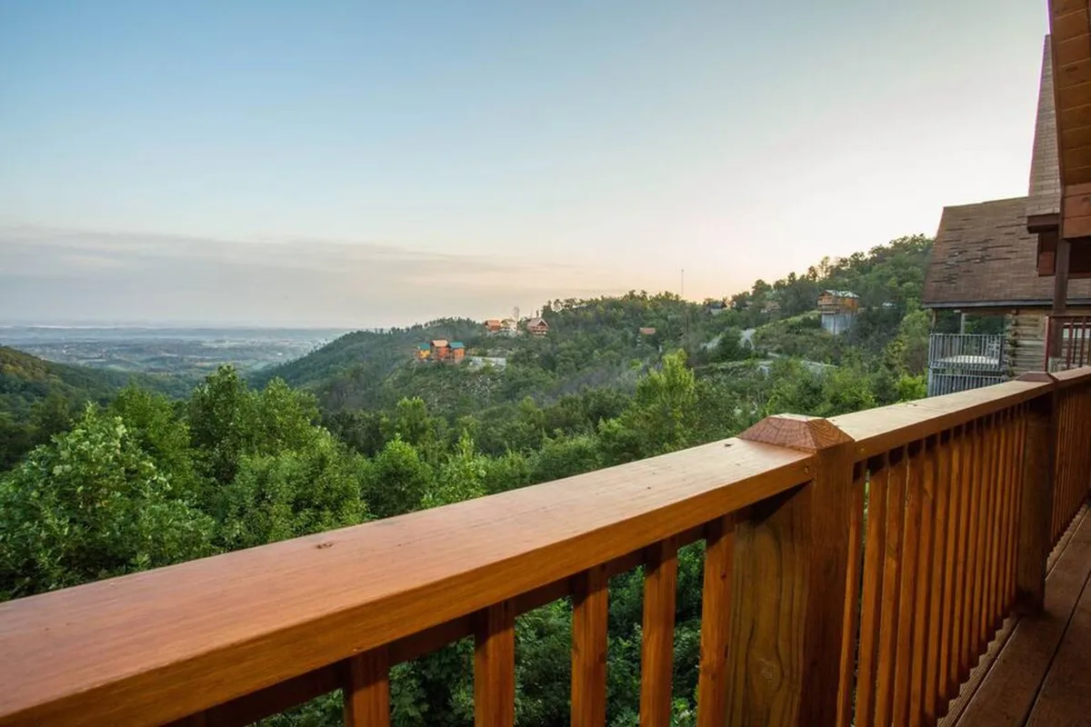 A wooden balcony overlooks lush green trees and distant mountain views, with nearby cabins visible. The balcony’s wooden railing and open air evoke a sense of relaxation and connection to the outdoors, inviting guests (and their pets) to enjoy the fresh mountain breeze after a day of adventures.