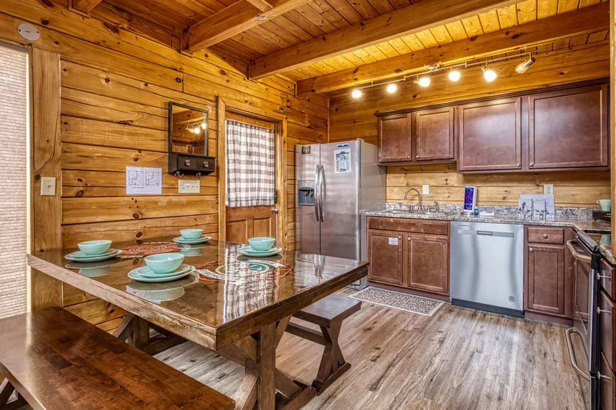 This image showcases a warm, inviting kitchen and dining area with a central wooden dining table, kitchen appliances, and rustic wooden cabinetry. The scene is set for family meals or casual gatherings, with the table ready to welcome guests.