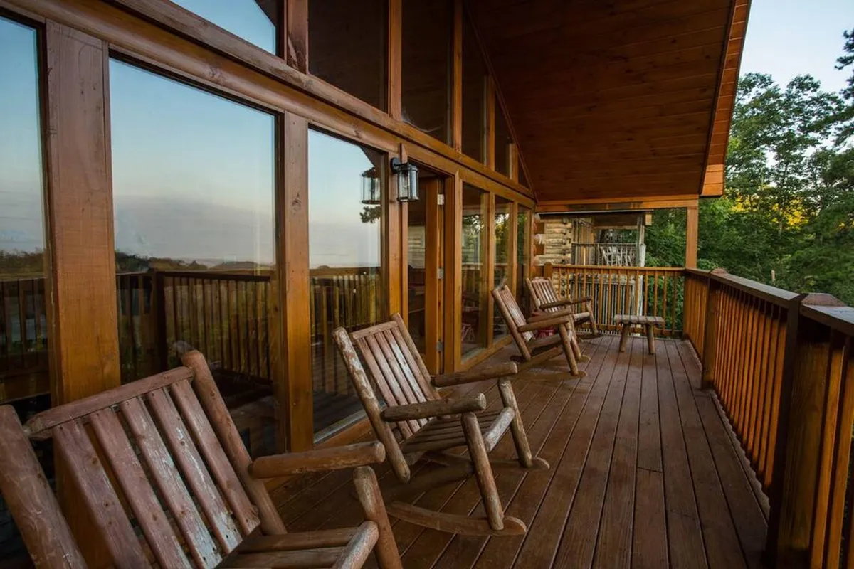 This image features inviting rocking chairs on a wooden deck with a railing, large glass windows, and lush trees surrounding the cabin. The setup suggests a comfortable place for friends to gather, socialize, and enjoy the mountain atmosphere together, with the natural landscape visible in the background.