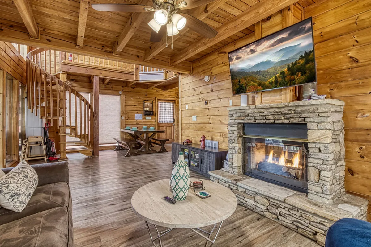 A cozy living room with a prominent stone fireplace, a spiral staircase, wooden beams on the ceiling, a large TV displaying a landscape image, and comfortable seating arrangements. Warm lighting and rustic decor create an inviting atmosphere, perfect for gathering.