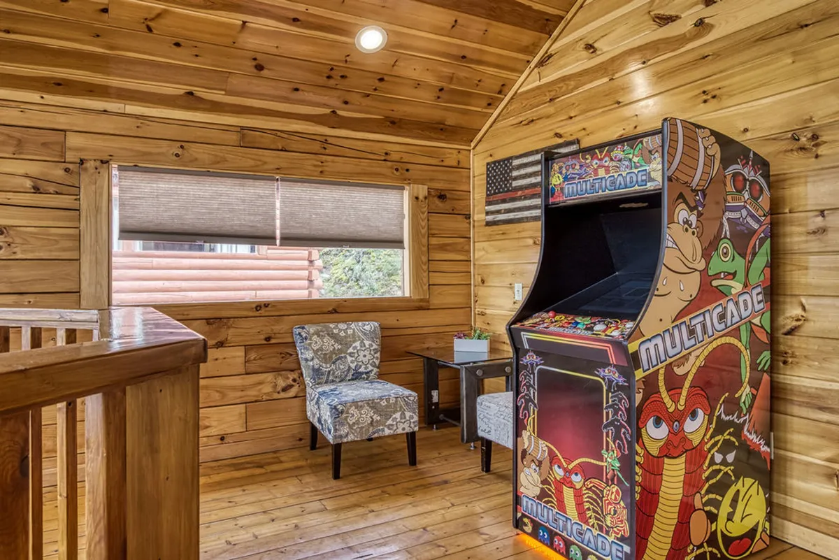 A cozy corner of the loft with a multicade arcade machine, warm wooden walls, and inviting seating. The arrangement suggests a nostalgic, fun spot for classic video games and relaxed gatherings.