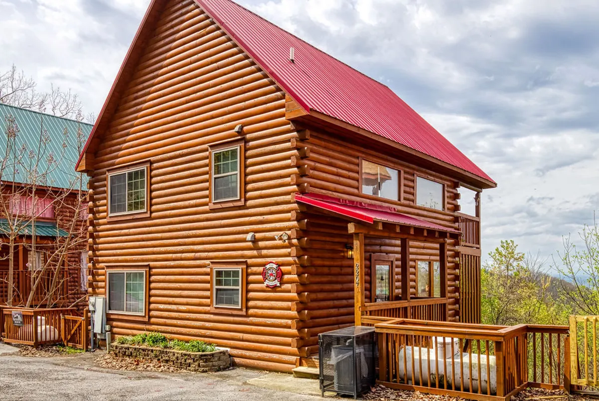 A log cabin with a distinctive red metal roof sits nestled among trees, with a spacious wooden deck inviting guests to relax outdoors. The setting exudes a cozy and welcoming vibe, perfect for family gatherings or simply unwinding after a day out. The natural surroundings suggest direct access to open spaces ideal for dog walks and playtime, as well as easy returns from nearby parks.