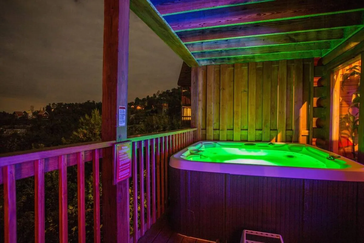 This exterior image shows a private hot tub set on a wooden deck with ambient lighting, surrounded by the serenity of the mountains. The view encompasses the natural setting and provides a sense of secluded luxury ideal for couples seeking relaxation and romance.