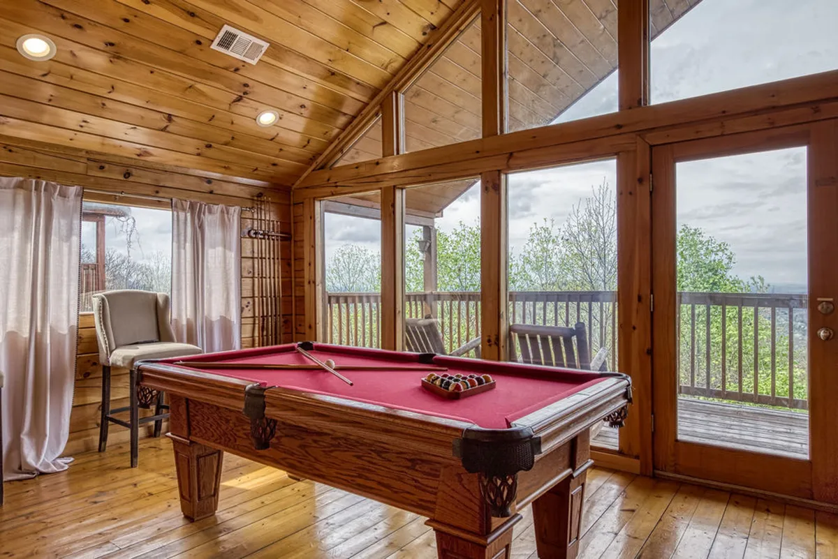 This interior image highlights a pool table by large windows with wooden flooring and a bar stool. The natural light and view from the windows blend indoor fun with the outdoor environment, creating an inviting space for friendly competition and group hangouts.