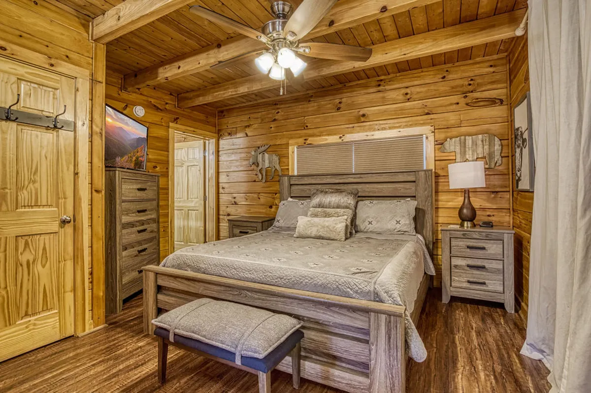 A tranquil bedroom with a wooden bed frame, nightstand and lamp, dresser, and wooden walls adorned with decorative animal art. The setting is peaceful and inviting, bathed in soft natural light and perfect for relaxation after a day in the mountains.